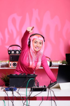 Téléchargez les photos : Artiste musical aux cheveux roses et chemisier s'amuser tout en jouant de la musique électronique à l'aide d'une console de mixage professionnelle, debout à la table de DJ dans le club la nuit. Asiatique femme appréciant performer - en image libre de droit