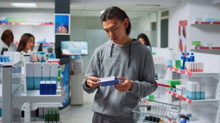 Foto de Hombre asiático comprobar botellas y cajas de pastillas en la farmacia, mirando medicina de la salud en los estantes. Examinar suministros farmacéuticos y vitaminas en paquetes, farmacia. - Imagen libre de derechos