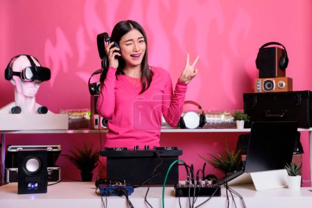 Photo for Artist with pink blouse wearing headset having fun while playing techno music using mixer console, standing at dj table in studio with pink background. Asian performer mixing electronic song in club - Royalty Free Image