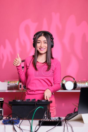 Téléchargez les photos : Parfumeur asiatique souriant debout à la table dj montrant signe d'approbation tout en exécutant de la musique électronique à l'aide d'une console de mixage professionnelle. Musicien debout en studio sur fond rose - en image libre de droit