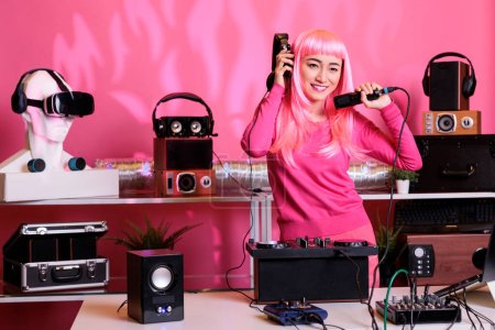 Photo for Asian musician standing at dj table enjoying to perform electronic music and talking with fans, using professional turntables in club at night. Performer with pink hair playing techno song - Royalty Free Image