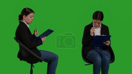 Photo for Confident businesswoman sitting on chair and analyzing papers, thinking about notes on clipboard files. Young manager acting thoughtful looking at official corporate documents, green screen. - Royalty Free Image