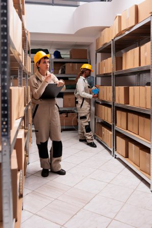 Photo for Warehouse manager in protective helmet counting boxes and writing on clipboard. Diverse stockroom employees working in storehouse, monitoring shipping process and packing parcels with adhesive tape - Royalty Free Image