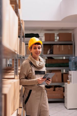 Photo for Smiling woman warehouse manager holding tablet and standing in delivery service storage portrait. Warehouse operator wearing protective helmet controlling shipment process - Royalty Free Image