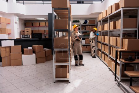 Photo for Warehouse operatives managing goods receiving inventory in stockroom. Young storehouse manager wearing protective helmet standing near tall shelf and writing on clipboard - Royalty Free Image