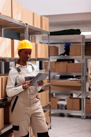 Photo for Warehouse operator using inventory app on digital tablet for goods management. Young african american storehouse manager wearing protective overall checking freight audit - Royalty Free Image