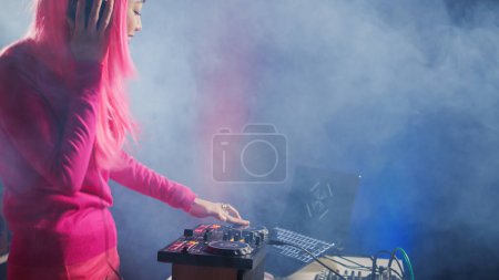 Téléchargez les photos : Artist creating musical performance with techno music using professional mixer console, having fun at night in club. Asian performer standing at dj table performing electronic song, celebrating album - en image libre de droit