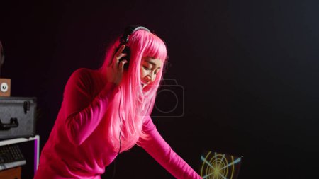 Téléchargez les photos : Cheerful artist working as dj playing song at turntables, mixing techno music with eletronic using audio equipment. Musician with pink hair having fun in performing in club at night time - en image libre de droit
