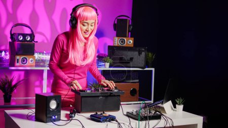 Téléchargez les photos : Smiling musician working as dj playing song at mixer console, mixing techno music with eletronic using audio equipment. Performer with pink hair having fun in performing in club at night time - en image libre de droit