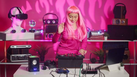 Téléchargez les photos : Asian performer dancing and interacting with fans in club at nightime, playing electronic remix at professional mixer console. Artist with pink hair mixing sounds using audio equipment - en image libre de droit
