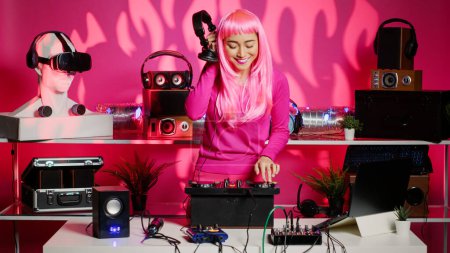 Photo for Happy artist mixing electronic remix at professional mixer console, listening music into headphones during party. Asian dj performer with pink hair dancing and having fun in club at night. - Royalty Free Image