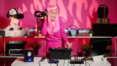 Téléchargez les photos : Happy artist with pink hair and blouse mixing sounds at professional mixer console, listening music into headset during party. Asian dj performer dancing and having fun in club at evening. - en image libre de droit