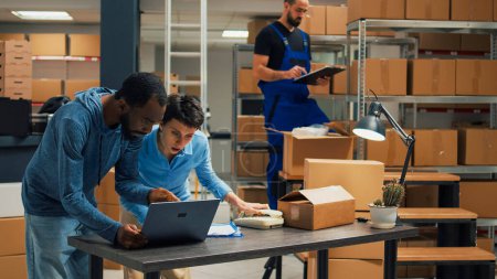 Foto de Young entrepreneurs doing teamwork to ship products in cardboard packages, checking quality of merchandise in storage room. Diverse people using laptop to plan goods shipment distribution. - Imagen libre de derechos