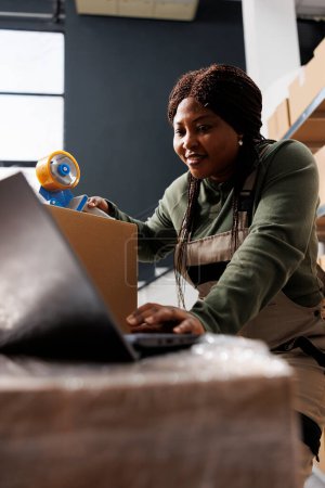 Photo for Stockroom worker analyzing online orders on laptop computer, preparing clients packages for shipping using cardboard boxes in storage room. African american employee working in warehouse - Royalty Free Image