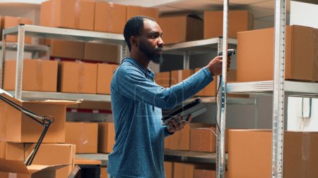 Foto de Male entrepreneur using scanner and tablet to check supplies inventory, working on products management. Young adult scanning merchandise in cardboard boxes on storehouse shelves. Handheld shot. - Imagen libre de derechos