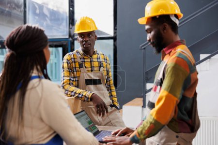 Photo for Warehouse managers checking pick ticket on laptop while talking in storage room. All black order pickers team standing in storehouse, chatting and preparing customer purchase for dispatching - Royalty Free Image