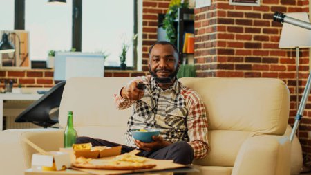 Photo for Modern male adult opening television to find film, eating snacks in bowl and drinking bottles of alcohol. Young smiling man watching favorite action movie, having multiple food. Tripod shot. - Royalty Free Image