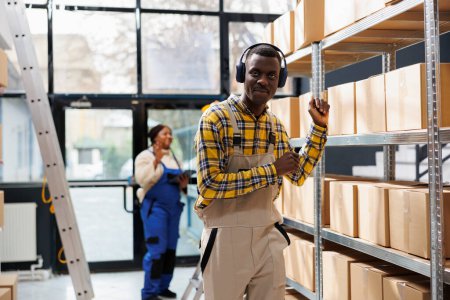 Photo for African american logistics manager dancing in factory storehouse and looking at camera. Young man wearing workwear listening to music and making hands movements in warehouse - Royalty Free Image