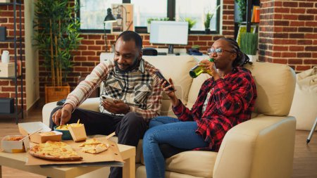 Foto de African american people using smartphones on couch, sitting together in front of television and browsing mobile phone apps. Modern partners enjoying leisure time with fast food meal. - Imagen libre de derechos