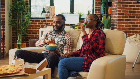Photo for African american couple having fun watching television, ordering burgers, pizza and nodles from delivery takeaway. Young partners enjoying movie or tv show, drinking bottles of beer. - Royalty Free Image