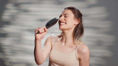 Téléchargez les photos : Happy flawless girl singing with hair brush on camera, fooling around and acting funny. Skincare model with glowing look having fun to promote self love and body positivity in studio. - en image libre de droit