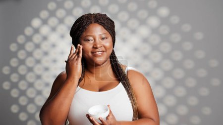 Photo for African american woman applying face cream on camera, beauty model advertising new skincare cosmetics. Beautiful girl using moisturizer or serum in studio, dermatology ad campaign. - Royalty Free Image