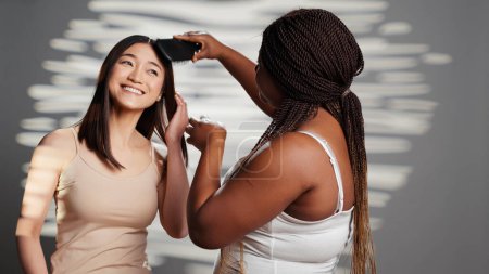 Téléchargez les photos : Belle femme brossant les cheveux de son amie à la caméra, posant pour promouvoir l'amitié et la féminité. Diverses femmes utilisant des cosmétiques et des produits de beauté publicitaires pour la campagne de soins du corps. - en image libre de droit