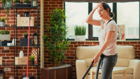 Téléchargez les photos : Casual girl using vacuum cleaner to tidy up apartment, cleaning dust and debris in living room. Young adult doing spring cleaning weekend activity chores, vacuuming wooden floors. - en image libre de droit