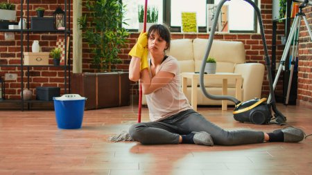 Photo for Caucasian housewife feeling sleepy after finishing spring cleaning session, sitting on clean wooden floor with mop and gloves. Tired woman being proud of tidy apartment, household chores. - Royalty Free Image