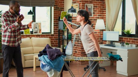 Téléchargez les photos : Young life partners having argument in living room, fighting over unfinished chores and spring cleaning. Aggressive woman being angry with husband while she irons clothes on ironing board. - en image libre de droit