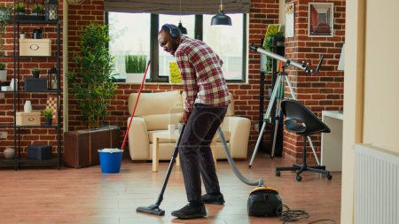 Téléchargez les photos : African american man dancing and vacuuming home floors, singing and listening to music on headset. Male person having fun spring cleaning, using vacuum to clean dust and dirt. - en image libre de droit