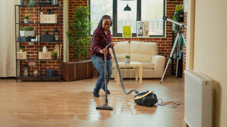 Téléchargez les photos : African american housewife using vacuum cleaner and listening to music on headset, enjoying modern spring cleaning at home. Young adult having fun with songs and vacuuming floors. - en image libre de droit