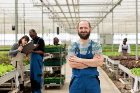 Photo for Smiling farm worker in green bio eco friendly agriculture organic hydroponic greenhouse with group of farmers. Locally grown chemical free vegetable crops using recycled water and non-GMO fertilizer - Royalty Free Image