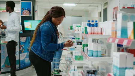 Photo for African american girl checking pharmaceutical products from pharmacy shelves, looking to buy medicaments and vitamins for healthcare. Young woman taking medicine and pills from drugstore. - Royalty Free Image