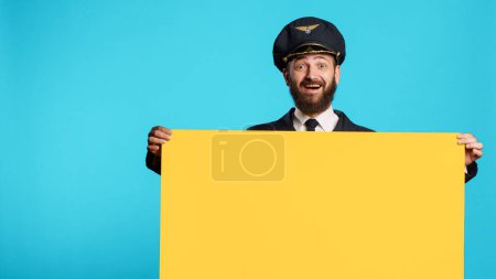 Foto de Cheerful commercial pilot showing empty mockup board on camera, working on advertisement with isolated copyspace billboard. Young smiling plane captain holding cardboard icon. - Imagen libre de derechos