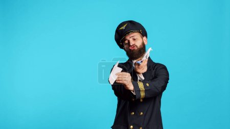 Foto de Airline pilot playing with paper and mini airplane, having fun with origami and small artificial aircraft in studio. Young male aviator wearing flying uniform, working on commercial flights. - Imagen libre de derechos