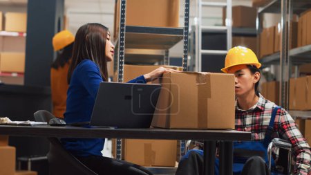 Téléchargez les photos : Asian man in wheelchair moving products in warehouse, checking merchandise from storage room racks. Male worker with disability preparing for order management, small business. Handheld shot. - en image libre de droit