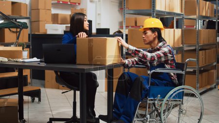 Téléchargez les photos : Asian wheelchair user working in warehouse space with supplies, man with physical disability carrying packs to check products before disribution. Working in disability friendly depot. - en image libre de droit