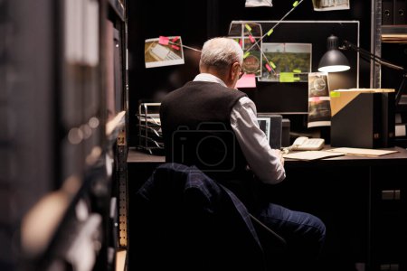 Photo for Elderly fbi investigator sitting at desk table in arhive room, analyzing crime evidence files. Private detective working overhours at mysterious suspect, checking victim report. Investigation concept - Royalty Free Image