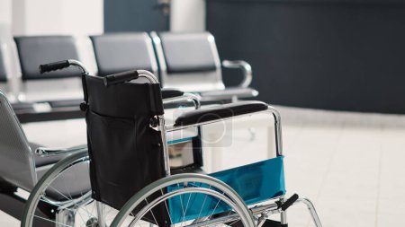 Photo for Wheelchair in empty waiting room used for physical recovery service on health care insurance, disability friendly clinic. Reception counter for registration with seats and medical forms. - Royalty Free Image