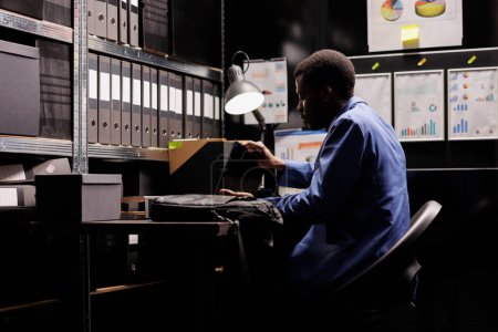 Téléchargez les photos : Le comptable afro-américain assis au bureau dans la salle des dossiers, travaillant à la recherche de dépositaires tard dans la nuit. Travailleur administratif analyse des preuves de surveillance, examen des dossiers d'enquêtes criminelles - en image libre de droit