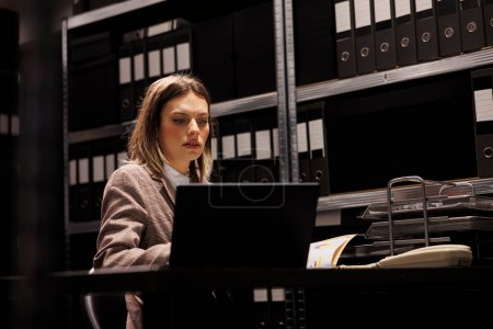 Photo for Caucasian bookkeeper reading administrative files, working late at night in corporate depository. Businesswoman in formal suit analyzing accountancy report, checking management research - Royalty Free Image
