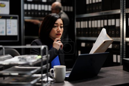 Photo for Bureaucratic administration employee checking accountancy budget plan data paperwork on laptop. Asian businesswoman executive in repository filled with document folders and graphical chart reports - Royalty Free Image