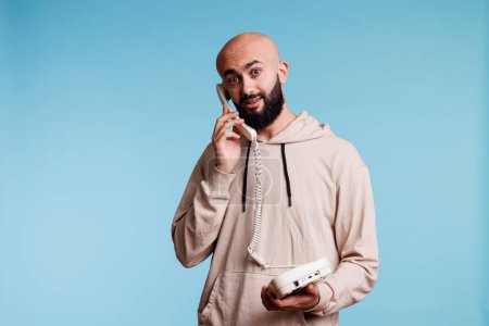 Photo for Excited arab man answering call while holding old fashioned landline phone portrait. Young person having conversation on retro telephone and looking at camera with amazed expression - Royalty Free Image