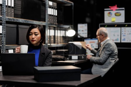 Photo for Accounting employees in consultancy bureaucratic workplace filled with business folders. Asian woman enjoying cup of coffee and senior coworker working in administrative file room office - Royalty Free Image