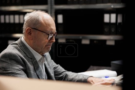 Photo for Elderly bookkeeping businessman at file cabinet office desk checking accounting analytical data paperwork. Administration senior executive in business company repository filled with document folders - Royalty Free Image