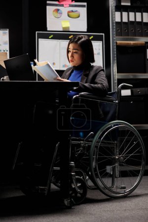 Téléchargez les photos : Comptable asiatique professionnel paralysé en fauteuil roulant vérifiant les documents administratifs dans la salle des dossiers. Femme d'affaires dans le bureau du classeur bureaucratique rempli de documents de facture et d'organigrammes - en image libre de droit