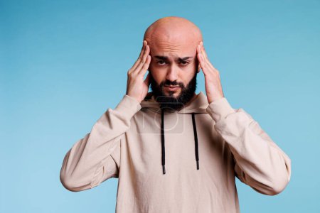 Photo for Exhausted arab man feeling tension in temples while suffering from headache studio portrait. Young bald bearded person having headache and looking at camera with pain grimace - Royalty Free Image