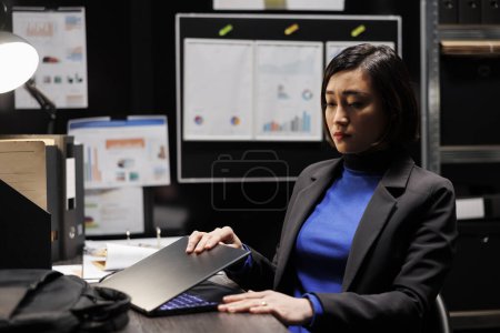 Téléchargez les photos : Femme d'affaires asiatique fermant ordinateur portable au travail se préparant à rentrer à la maison. Comptabilité consultant exécutif entouré de dossiers comptables bureau statistiques organigrammes de données - en image libre de droit