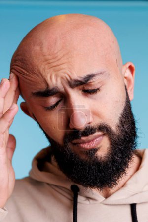 Téléchargez les photos : Homme arabe avec les yeux fermés ayant la migraine et se frottant la tête dans la douleur. Jeune adulte chauve barbu souffrant de maux de tête et tenant temple tout en posant sur fond bleu - en image libre de droit